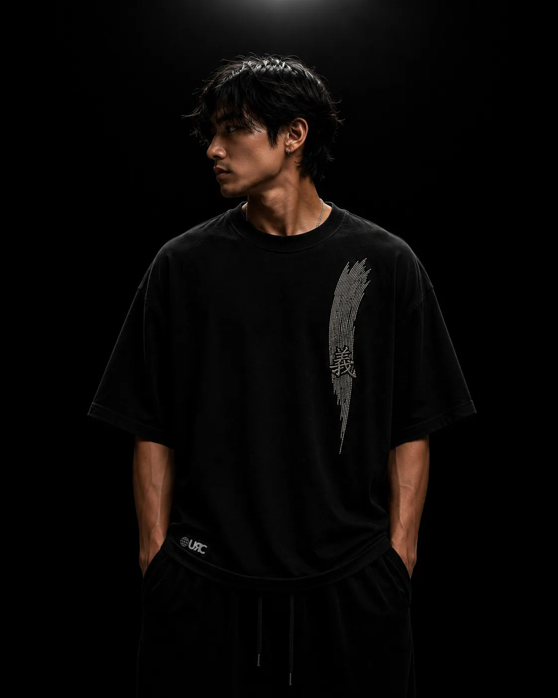 Solitary figure wearing the oversized black GI tee, showcasing the engineered drop-shoulder silhouette and stark raised satin chest embroidery.