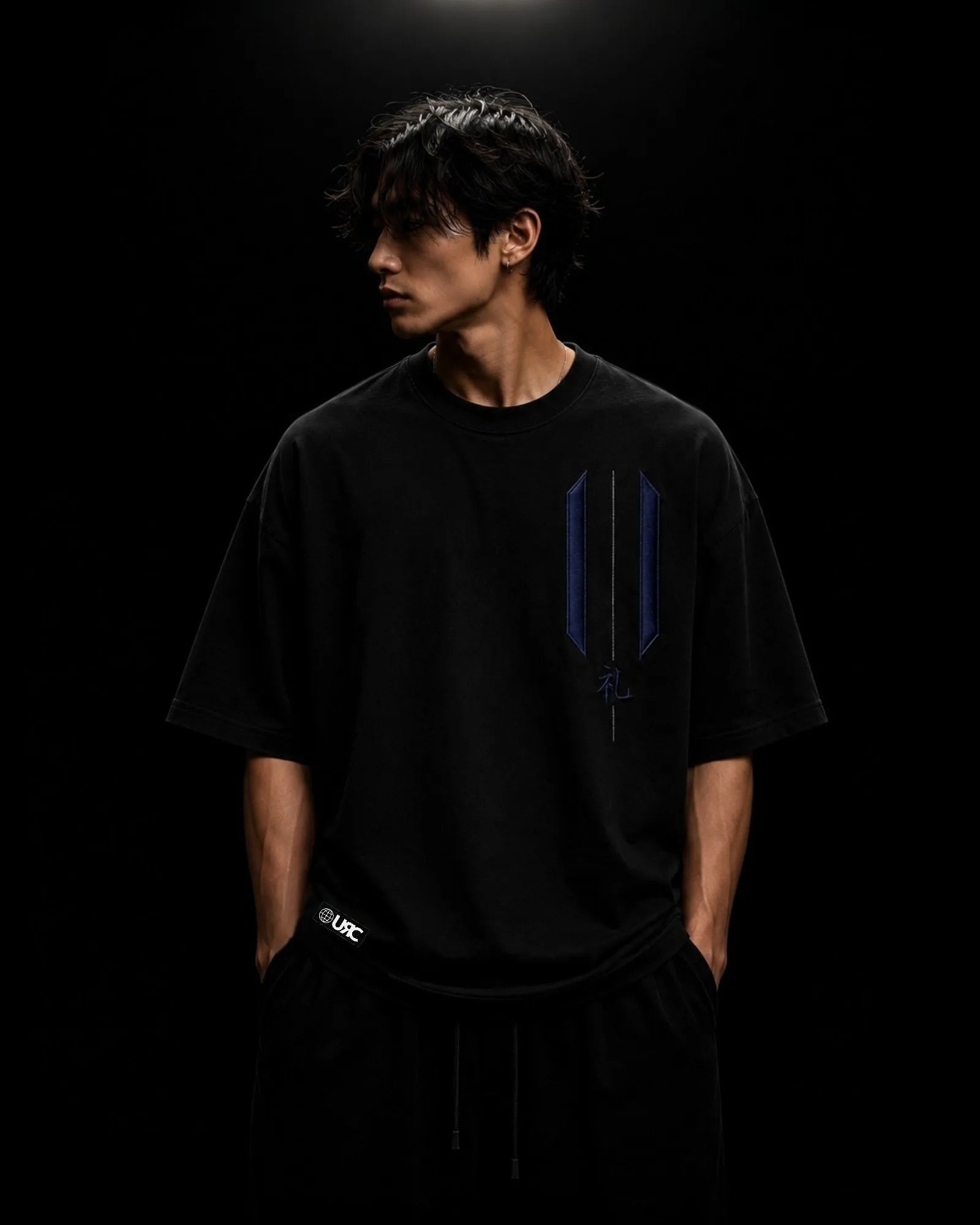 Solitary figure wearing the oversized black REI artifact, highlighting the engineered drop-shoulder silhouette and the minimal ceremonial embroidery positioned over the heart, where respect begins before action.