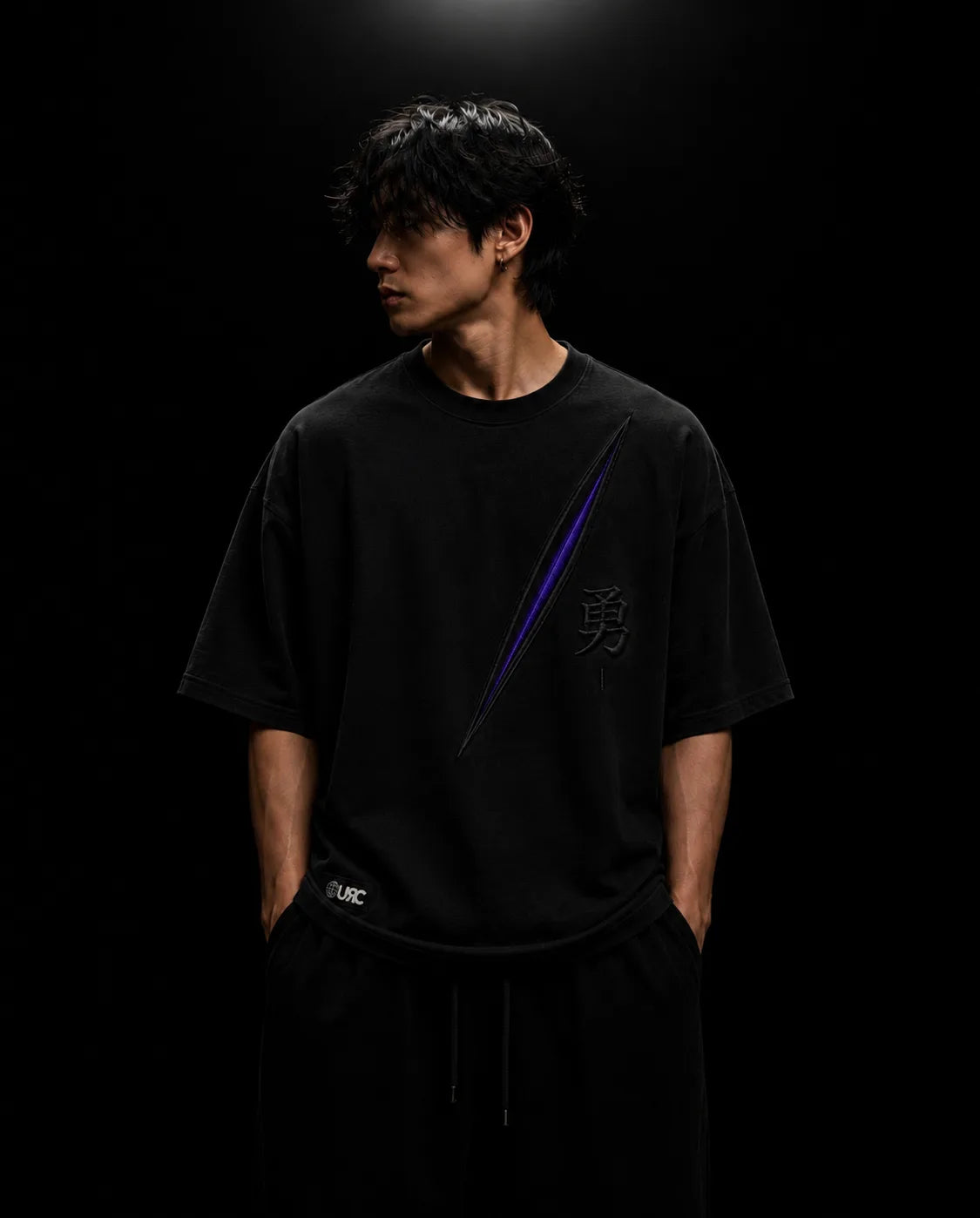 Solitary figure wearing the oversized black YU artifact, showcasing the engineered drop-shoulder architecture and striking raised satin chest embroidery.
